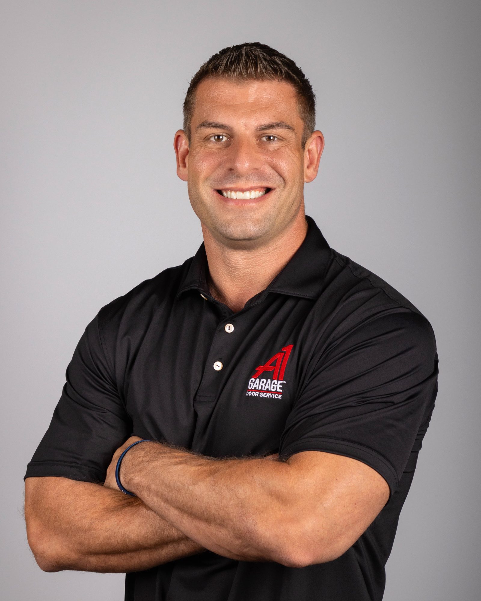 Tommy Mello, a man with short dark hair, smiling with his arms crossed, wearing a black polo shirt with the red A1 logo